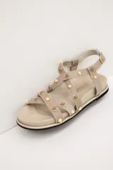 Sandalias de cuero color beige, con tiras cruzadas en el empeine decoradas con tachas y esferas doradas, base anatómica y pulsera ajustable al tobillo.