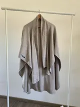 Ruana de tela tipo tapiz con textura, en color gris. Presenta un diseño abierto con caída estructurada y etiqueta decorativa en el lateral.