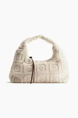 Cartera de mano tejida en crochet con motivo de cuadrados, color crudo, con asa corta integrada y tiras de cuero marrón como detalle de cierre.