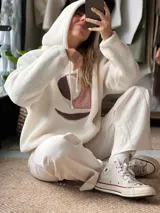 Buzo tipo hoodie de corderito color manteca, con capucha y cordones ajustables. Presenta un diseño abstracto frontal con apliques de textura suave en tonos rosa viejo y marrón.
