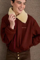Campera bomber marrón de paño de lana, con cierre frontal, cuello de corderito sintético blanco desmontable, bolsillos laterales, puños y dobladillo elásticos, y logo bordado en el pecho.