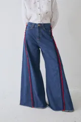 Jean de corte wide leg en color azul denim, con detalle de apliques de crochet en color rojo y azul a lo largo de las costuras laterales frontales. Presenta ruedo deshilachado y cierre frontal con botón metálico.