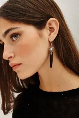 Aro de tipo gancho francés con colgante de cuerno de hueso y detalle superior de piedras y cristales facetados en tonos neutros. Presenta un diseño de inspiración étnica.