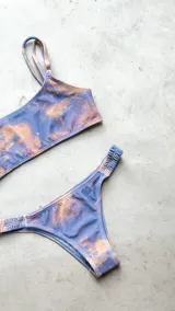 Conjunto de bikini con corpiño estilo bandó alto y breteles regulables, con lazo para atar en la espalda. La bombacha es vedetina con frunce elástico en los costados. Presenta un estampado tipo tie-dye o marmolado en tonos azules y naranjas/terracota.