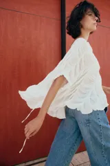 Top blanco de cuello redondo y mangas tipo capa, con textura fruncida, bajo asimétrico, lazos decorativos en el escote y en los laterales, y cierre posterior con botón.