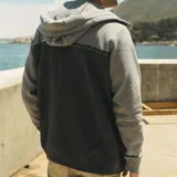 Campera canguro Maui and Sons de felpa con cierre completo y capucha ajustable con cordón. Presenta un diseño bicolor en tonos gris claro y gris oscuro, separado por una franja horizontal con patrón de olas y detalles en turquesa. Cuenta con bolsillos laterales tipo ojal y puños y ruedo de rib elástico.