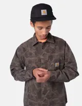 Camisa de manga larga Carhartt con estampado camuflado en tonos verdes y marrones. Presenta cuello clásico, cierre frontal con botones, un bolsillo en el pecho con etiqueta de la marca y puños abotonados.
