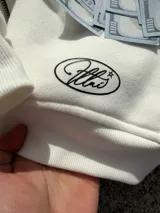 Campera canguro blanca con cierre frontal, capucha y bolsillos tipo kankaro. Presenta un estampado frontal con un óvalo con texto cursiva rodeado de estrellas negras.