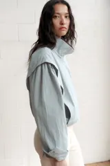 Chaqueta corta de cuello subido con manga larga, cierre frontal con cremallera y botones a presión ocultos. Presenta bolsillos delanteros y puños con pliegues.