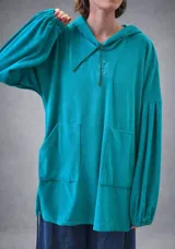 Buzo tipo canguro oversize de tela plush, color azul claro. Presenta capucha, cierre frontal parcial con cordón turquesa y bolsillos canguro frontales. Las mangas son largas y abullonadas con puños ajustados.