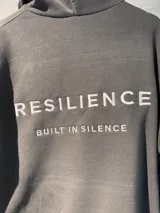 Canguro con capucha de algodón con felpa, color gris, con diseño bordado en la espalda que incluye el texto 'RESILIENCE' y 'BUILT IN SILENCE'.