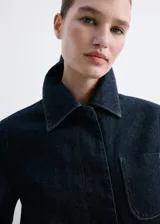 Chaqueta corta de denim en color azul oscuro, con cuello clásico, cierre frontal con botones, mangas largas y un bolsillo aplicado en el pecho.