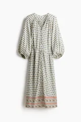 Vestido túnica largo oversize de color beige con estampado geométrico azul y detalles en el borde inferior. Presenta escote en V con cordones finos, mangas globo abullonadas de longitud 3/4 con puños elásticos y cordón ajustable en la cintura.