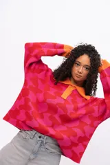 Buzo de tejido de punto con diseño geométrico en tonos fucsia y rojo, cuello tipo polo con cierre de botones y puños en color naranja contrastante. Presenta un corte holgado y oversize.