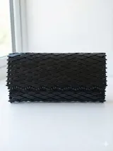 Cartera tipo sobre de mano color negro, con diseño estructurado formado por cuentas o piezas de madera formando un patrón texturizado similar a escamas.