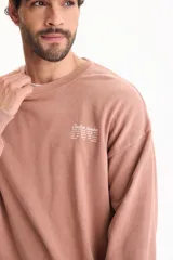 Buzo deportivo sin friza color marrón claro, con cuello redondo, mangas largas, puños y cintura elásticos, y pequeño texto estampado en el pecho.