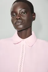 Camisa de popelina color rosado, con cuello redondo amplio, manga corta abullonada y detalle de bordado decorativo con perlas en la botonera frontal oculta.