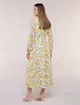 Vestido maxi de gasa con estampado floral en tonos amarillos y verdes sobre base blanca, escote en V cruzado, mangas largas y cintura elastizada con lazo.