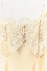 Vestido midi de estilo lencero confeccionado en viscosa satinada color crema. Presenta escote recto con tirantes finos, detalle de encaje floral en el pecho y el ruedo, y pliegues verticales en el frente.