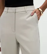 Pantalón de sastrería marca Cortelle, de corte recto y color beige. Presenta pliegues delanteros, cintura con trabillas, cierre frontal y bolsillos traseros embutidos.