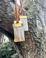 Collar corto con dije rectangular de nácar en tonos marrones y beige, con detalle metálico dorado en la parte superior y tiento de cuero marrón.