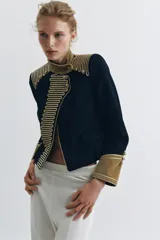 Chaqueta de estilo militar en color negro, con cuello subido y puños en terciopelo color verde oliva. Presenta detalles de alamares dorados en el frente y hombros, bolsillos con solapa y cierre frontal con botones metálicos en relieve.