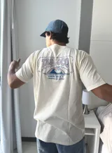 Remera de manga corta color beige jaspeado, con un estampado gráfico en la espalda que incluye el texto "TAS LOCO" y "SUNNY WITH A CHANCE OF STOKE" con un diseño de una persona en el agua con una ballena.