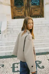 Chaqueta corta estilo trench color marrón oscuro, de corte cruzado con doble botonadura, cuello solapa y hombreras. Presenta mangas largas con puños anchos y bolsillos delanteros de vivo.