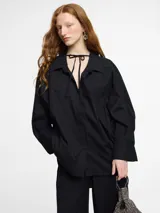 Camisa negra de corte oversize y manga larga con puños abotonados. Presenta un cuello clásico con un lazo fino para anudar en la parte superior y un diseño con bajo asimétrico, siendo más larga en la parte posterior.