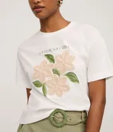 Remera de algodón color blanco, con cuello redondo y manga corta. Presenta un estampado con la frase 'Soy flor. Soy latina' y flores bordadas en relieve con textura tejida.