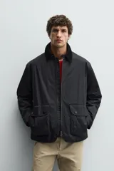 Parka de corte relajado en color negro, confeccionada en tejido de algodón con interior ligeramente acolchado. Presenta cuello de solapa en pana, cierre frontal con cremallera y bolsillos delanteros tipo plastrón con solapa y botones a presión.