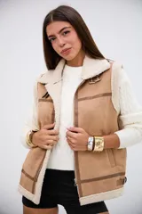 Chaleco de gamuza sintética color camel con interior y bordes de corderito sintético color crema. Presenta cierre frontal metálico, detalles de tiras con hebillas en el cuello y en la parte inferior, y costuras reforzadas con ribetes de cuero sintético.