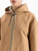 Chaqueta color camel con capucha, cierre frontal y bolsillos laterales. Presenta un diseño de corte recto y forro interior a cuadros, con detalle de bordado en el pecho.