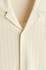 Camisa de manga corta con cuello abierto, confeccionada en tejido con textura. Presenta un corte holgado y bajo recto con aberturas laterales.