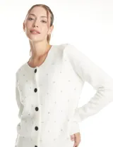 Cardigan de punto color blanco con cuello redondo, manga larga y cierre frontal con botones forrados en terciopelo negro. Presenta un diseño de textura acolchada con apliques de strass distribuidos por toda la prenda.