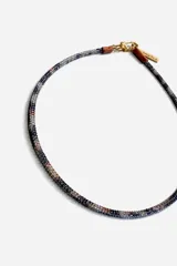 Gargantilla de cuentas tejidas con patrón de camuflaje en tonos azul, blanco y cobre. Cuenta con cierre de mosquetón metálico dorado y una pequeña placa con logo grabado.