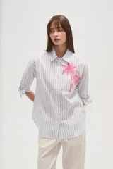 Camisa de algodón con rayas verticales azules y blancas, corte ancho y manga larga con lazos ajustables en los puños. Presenta un bordado de dos palmeras en color rosa brillante en el área del pecho.