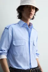 Camisa de algodón color celeste, corte relaxed fit, con cuello solapa, manga larga y trabillas en los hombros. Presenta dos bolsillos de plastrón con solapa en el pecho.