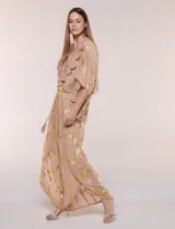 Pantalón palazzo confeccionado en gasa transparente color rosa con diseño estampado metalizado dorado. Presenta cintura elástica con cordón ajustable con borlas, piernas anchas y diseño cruzado al frente con aberturas laterales y detalles de puntillas.