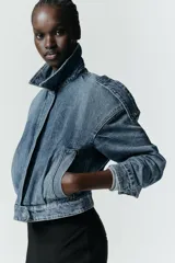 Cazadora de denim estilo crop con cuello solapa, manga larga y cierre frontal con cremallera y botón metálico. Presenta bolsillos de vivo en el delantero y detalle de trabillas en los hombros.