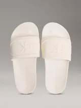 Ojotas tipo slide de color blanco hueso, con tira ancha acolchada y monograma de CK en relieve. La suela es de espuma.