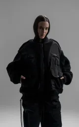 Campera estilo bomber unisex, confeccionada en una combinación de corderito sintético y paneles de ripstop. Presenta un diseño utilitario con múltiples bolsillos frontales con cierres y broches a presión, cuello, puños y ruedo en ribb elástico.