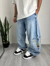 Pantalón de jean estilo baggy, color azul claro con efecto lavado. Presenta un diseño holgado, costuras reforzadas en las rodillas y un bolsillo lateral tipo cargo con un estampado gráfico de una cadena dorada y un candado con calavera.