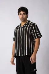 Camisa de manga corta tejida al crochet con rayas verticales negras sobre fondo crudo. Presenta cuello clásico con solapa y cierre frontal de botones.
