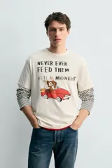 Camiseta de corte relajado color crudo, con cuello redondo y manga corta. Presenta un estampado frontal con una ilustración de Gizmo de la película Gremlins conduciendo un auto rojo y el texto 'Never ever feed them after midnight'.