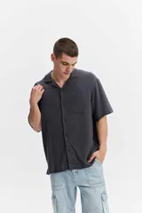 Camisa de manga corta color gris oscuro, confeccionada en muselina de algodón, con cuello abierto, cierre frontal de botones y un bolsillo aplicado en el pecho.