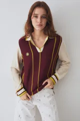 Remera tejida de punto estilo polo con mangas largas en color crema y cuerpo en color bordó con rayas verticales amarillas. Presenta cuello tipo polo, aberturas laterales y un pequeño parche circular con diseño de sol en el pecho.