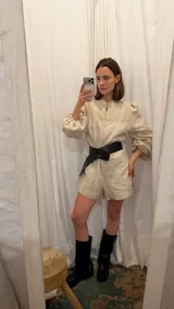 Mono corto de lino color beige, con cuello camisero, cierre frontal, mangas largas abullonadas y cinto ajustable en la cintura.