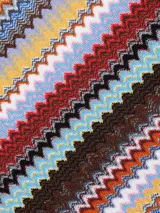 Chal tipo bufanda de tacto suave con patrón de zigzag multicolor en franjas verticales. Presenta flecos en los bordes.