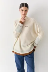 Buzo tejido de punto con cuello alto, diseño holgado y aberturas laterales. Presenta un cuerpo en color azul melange con detalles de franjas en color crema y verde en los puños y el ruedo. Incluye un pequeño bordado en el pecho.